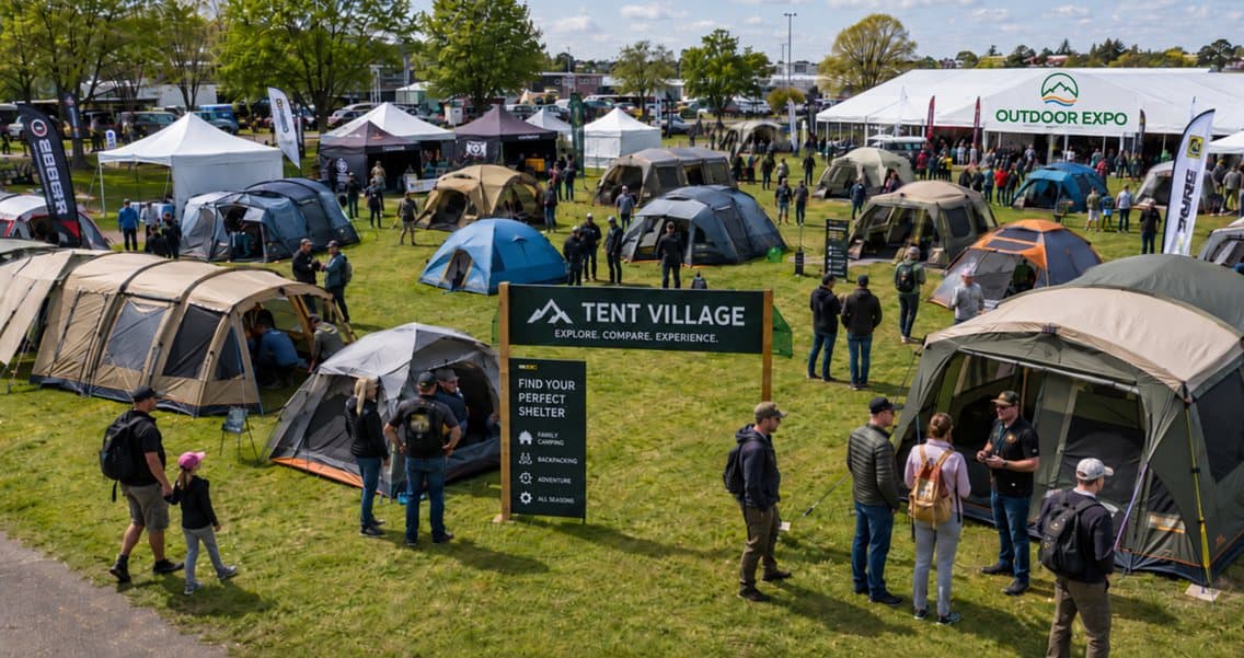 Family tents pitched and open around the Tent Village sign at Outdoor Expo, with visitors walking between them