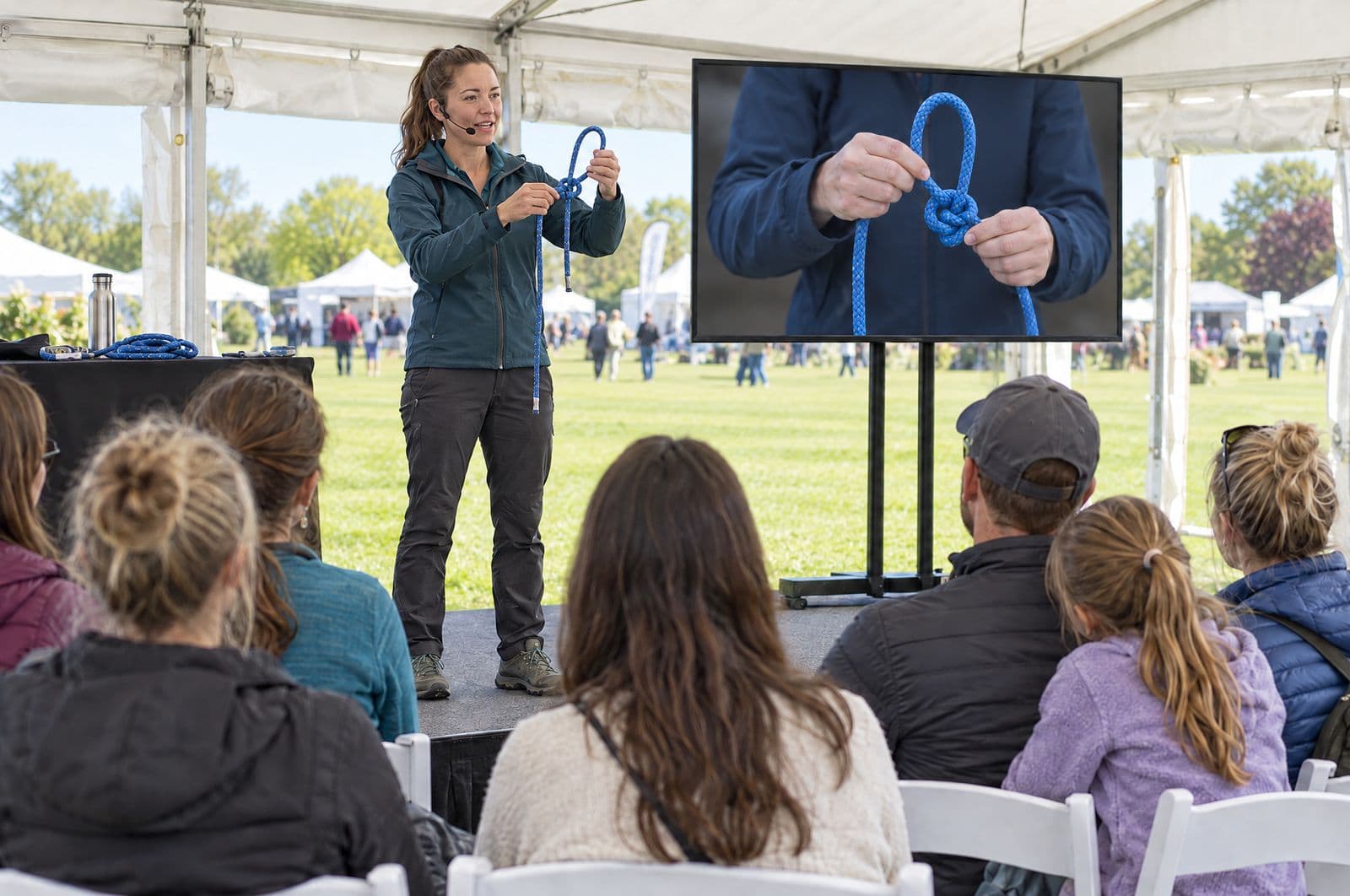 A presenter on the Demo Tent stage shows how to tie a bowline while a big screen beside her shows her hands up close